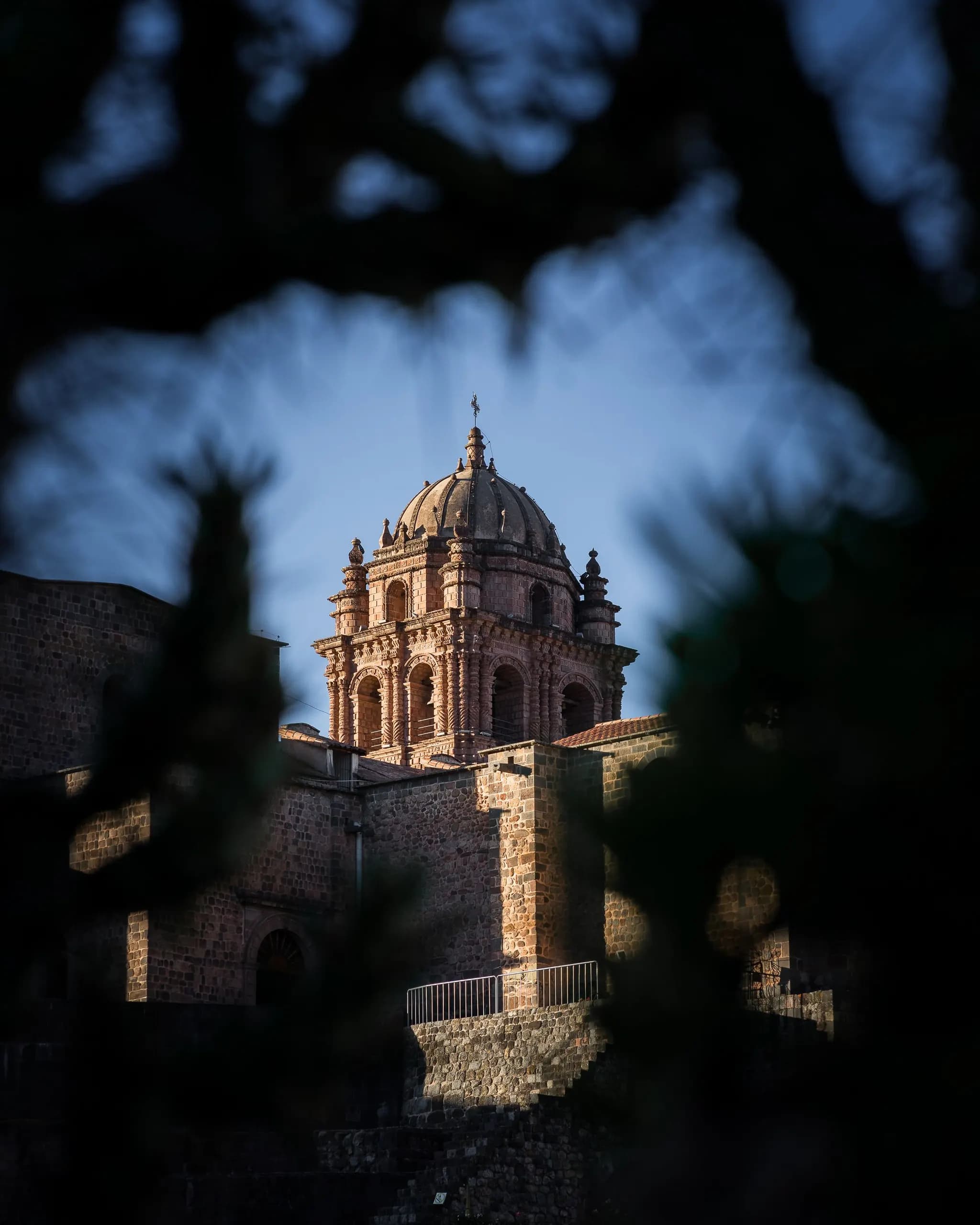 © jair romero · Pexels Sunlit stone bell tower of a church in Cusco, framed by tree branches