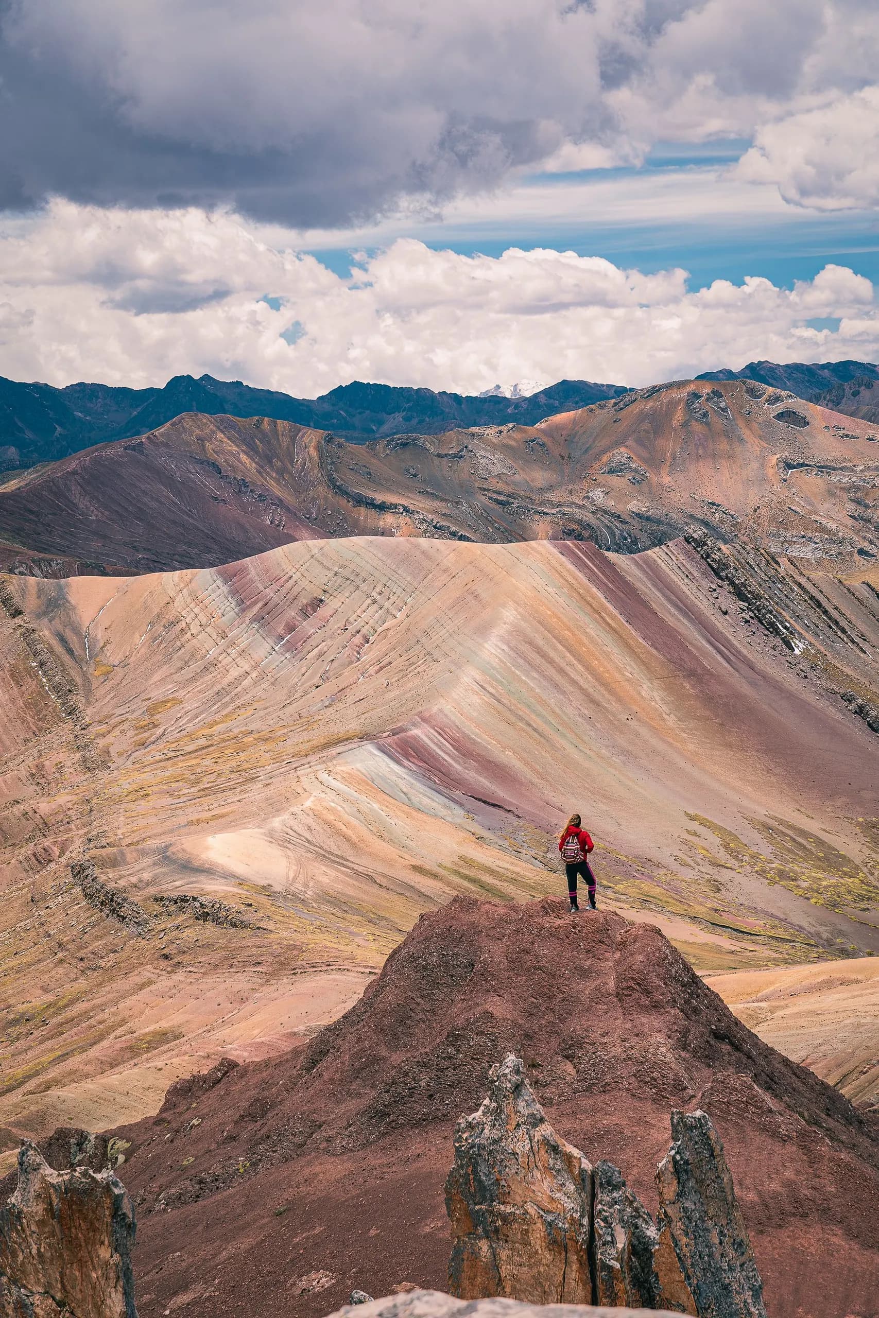 © jose galarza · Pexels Hiker standing on a rocky viewpoint facing the slopes of Rainbow Mountain beneath a cloudy sky